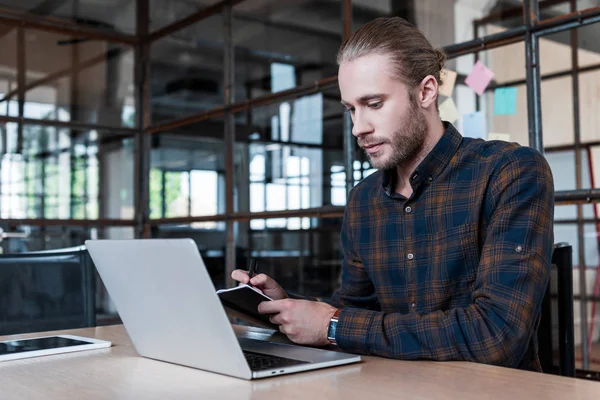 Beau jeune homme d'affaires prenant des notes et travaillant avec ordinateur portable dans le bureau — Photo de stock