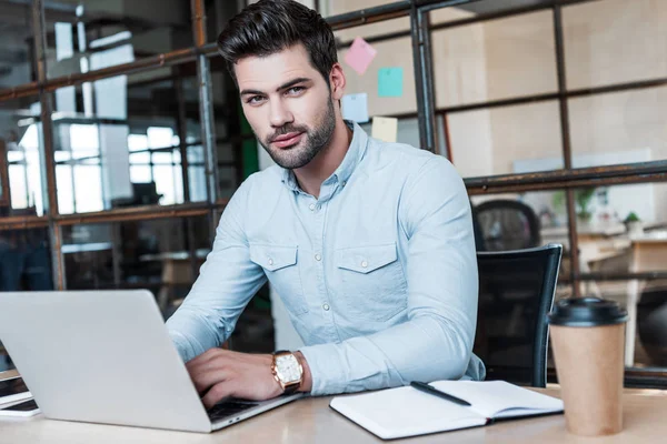 Handsome young businessman using laptop and looking at camera at workplace — Stock Photo