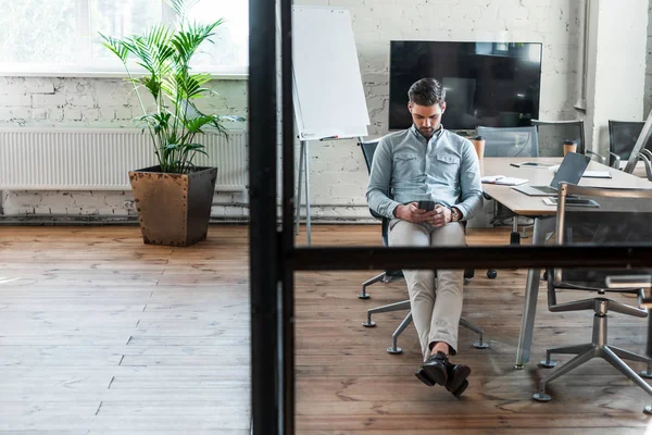 Bonito jovem empresário usando smartphone enquanto sentado no local de trabalho — Fotografia de Stock