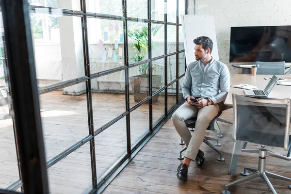 Visão de alto ângulo do jovem empresário usando smartphone e olhando para longe enquanto sentado no escritório — Fotografia de Stock