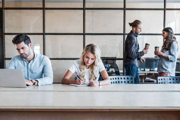 Junge Geschäftsfrau und Geschäftsfrau, die am Arbeitsplatz arbeitet, während ihre Kollegen im Hintergrund Kaffee trinken — Stockfoto