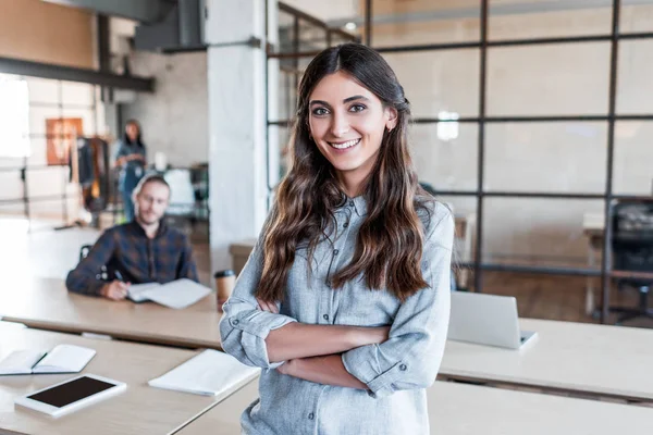 Beautiful young businesswoman standing with crossed arms and smiling at camera in office — Stock Photo