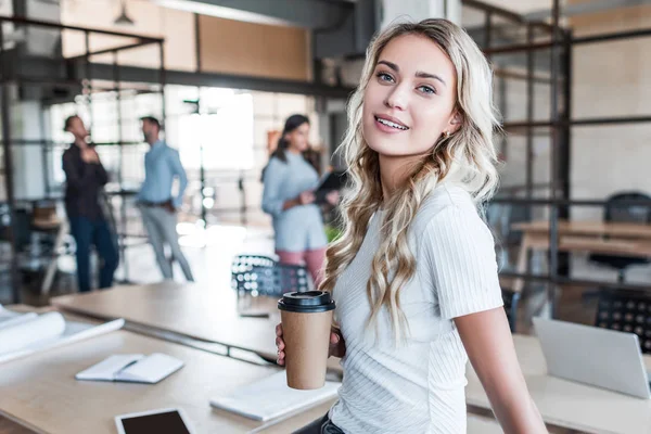 Atractiva joven empresaria sosteniendo taza de papel y sonriendo a la cámara en la oficina - foto de stock