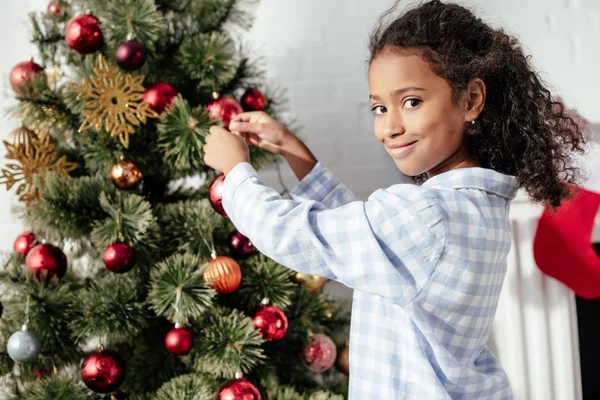 Adorable african american child in pajamas decorating christmas tree with baubles and looking at camera at home — Stock Photo
