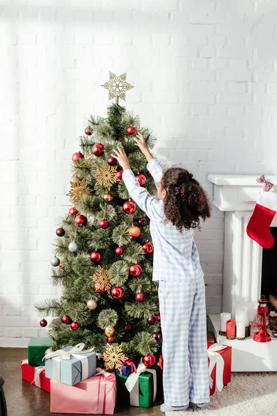 Visão traseira da criança americana africana em pijama decorando árvore de natal com bugigangas em casa — Fotografia de Stock