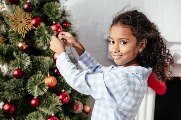 Adorable niño afroamericano sonriente en pijama decorando el árbol de navidad con adornos y mirando a la cámara en casa - foto de stock