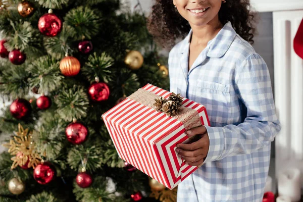 Обрезанный образ улыбающегося африканского американского ребенка в пижаме с рождественским подарком дома — стоковое фото