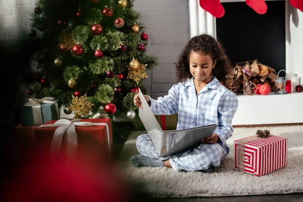 Foco seletivo da adorável criança afro-americana em pijamas livro de leitura perto da árvore de natal em casa — Fotografia de Stock