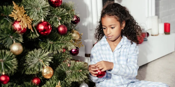 Adorável criança afro-americana em pijama decorando árvore de natal com bolas vermelhas em casa — Fotografia de Stock