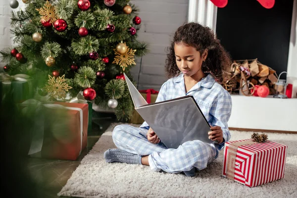 Adorable african american child in pajamas reading book near christmas tree at home — Stock Photo
