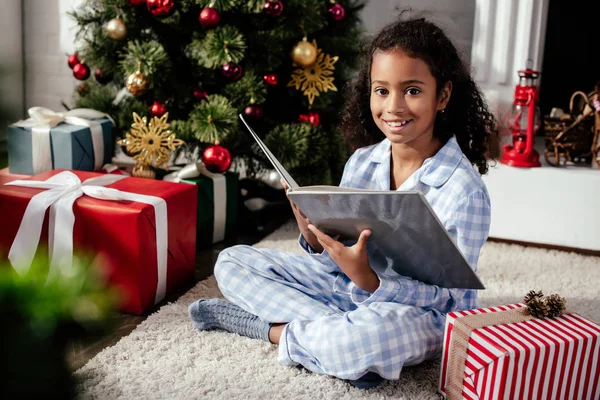 Niño afroamericano adorable sonriente en pijama sosteniendo libro cerca del árbol de Navidad y mirando a la cámara en casa - foto de stock