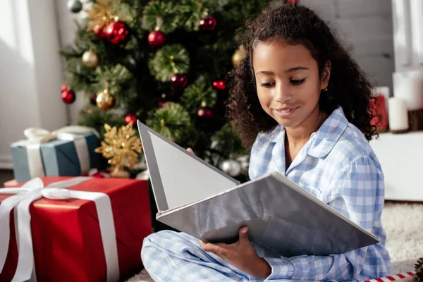 Alegre adorable afroamericano niño en pijama lectura libro cerca de árbol de Navidad en casa - foto de stock