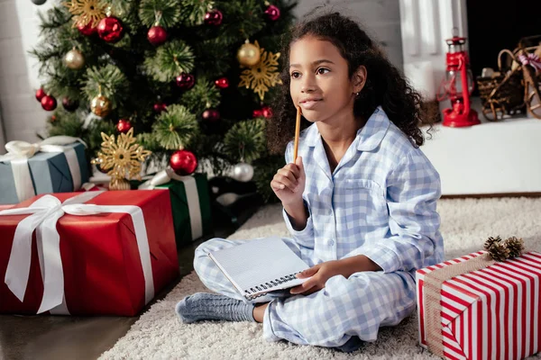 Adorable niño afroamericano pensativo en pijama sentado con copybook cerca del árbol de Navidad en casa - foto de stock