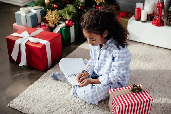 Adorável criança afro-americana em pijama escrevendo algo para copybook em casa, conceito de natal — Fotografia de Stock