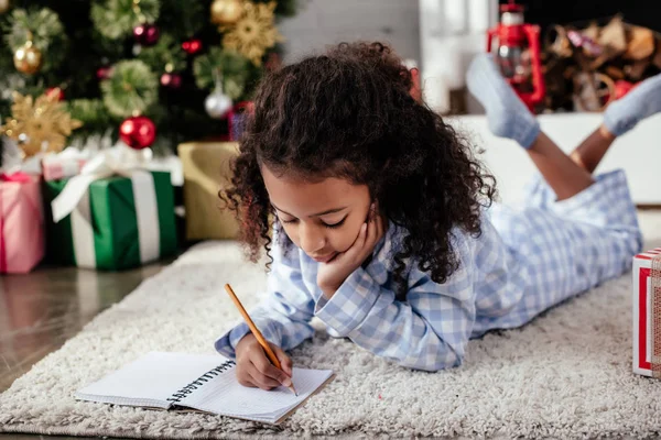 Adorable african american child in pajamas lying on floor and writing something to copybook at home, christmas concept — Stock Photo
