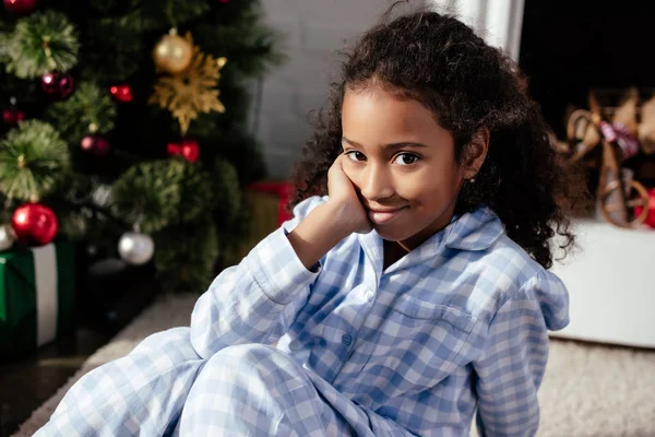 Adorable afroamericano niño en pijama sentado en el suelo y mirando a la cámara en casa, concepto de Navidad - foto de stock