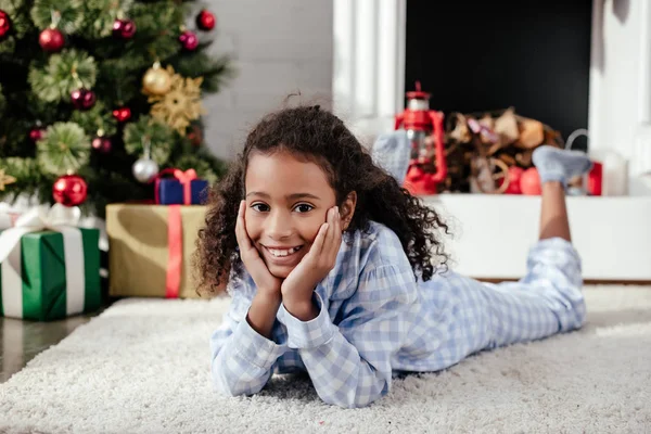 Adorável criança afro-americana de pijama deitada no chão e olhando para a câmera em casa, conceito de natal — Fotografia de Stock