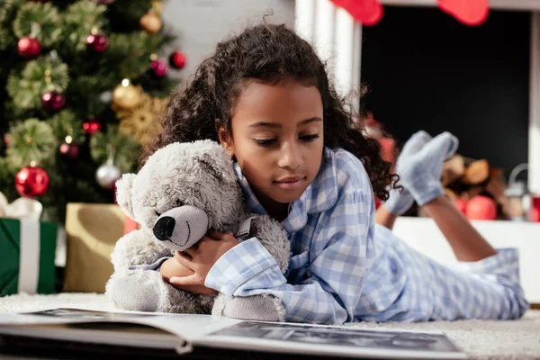 Adorable afroamericano niño en pijama con osito de peluche mirando álbum de fotos en el suelo en casa - foto de stock