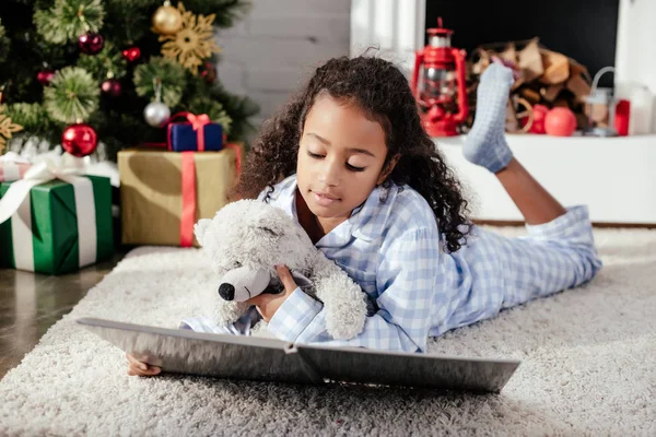 Adorable african american child in pajamas with teddy bear reading book on floor at home, christmas concept — Stock Photo