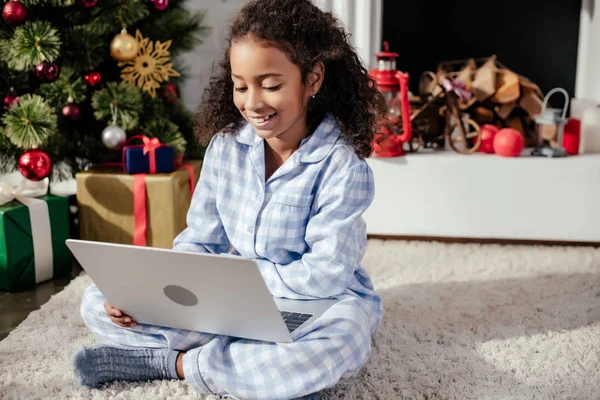 Sorridente adorabile bambino africano americano in pigiama utilizzando il computer portatile a casa, concetto di Natale — Foto stock