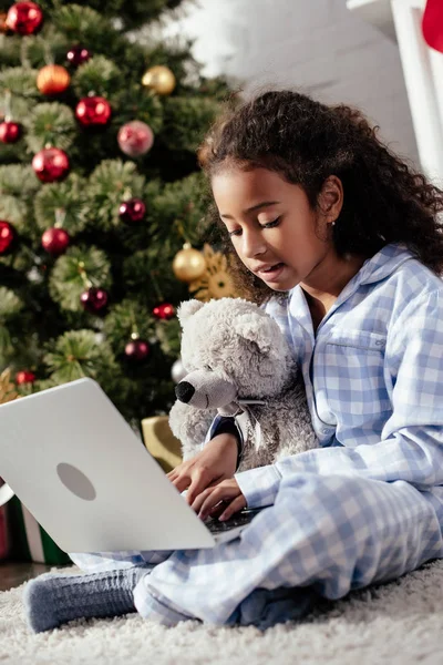 Чарівна афроамериканська дитина в піжамі з плюшевим ведмедем використовує ноутбук вдома, різдвяна концепція — стокове фото