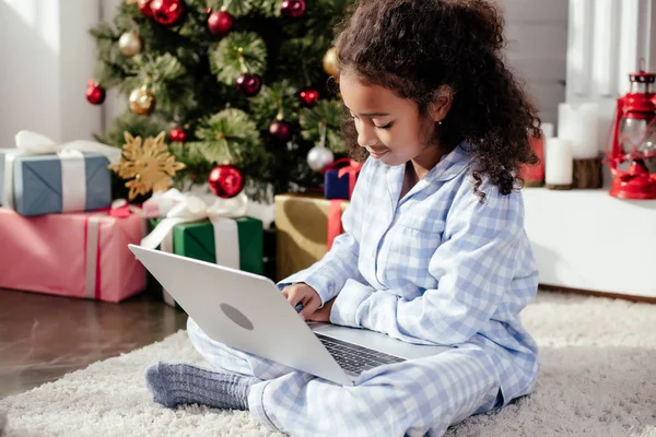 Adorable african american child in pajamas using laptop at home, christmas concept — Stock Photo