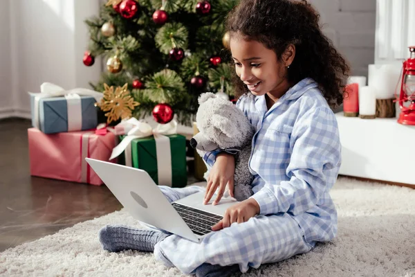 Criança americana africana adorável feliz em pijama com ursinho de pelúcia usando laptop em casa, conceito de natal — Fotografia de Stock
