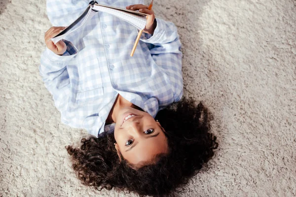 Top view of cheerful adorable african american child in pajamas lying on carpet with copybook in room — Stock Photo
