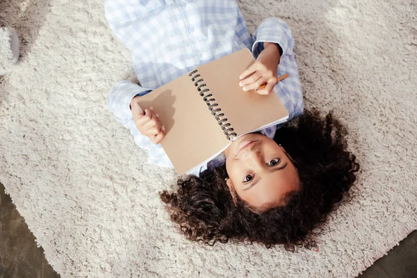 Top view of adorable african american child in pajamas lying on carpet with copybook and looking at camera — Stock Photo