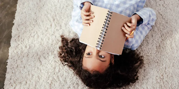 Top view of adorable african american child in pajamas lying on carpet and covering face with copybook in room — Stock Photo