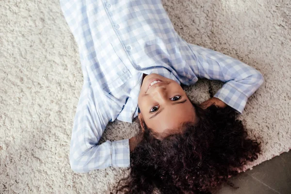Вид сверху на улыбающегося очаровательного африканского ребенка в пижаме лежащего на ковре в комнате — стоковое фото