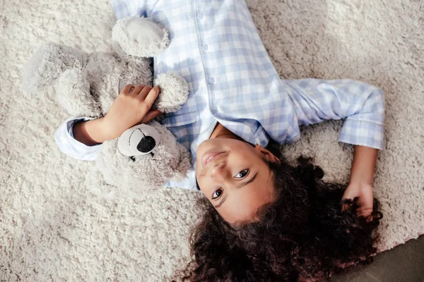 Vista superior de adorable niño afroamericano en pijama acostado con oso de peluche en la alfombra en la habitación y mirando a la cámara - foto de stock