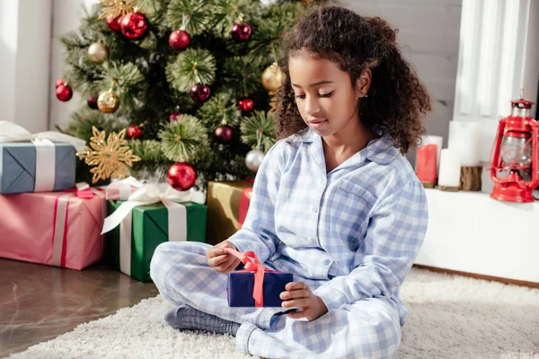 Adorable african american child in pajamas opening christmas present near christmas tree at home — Stock Photo