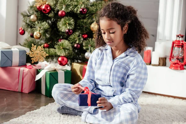 Adorável criança afro-americana em pijama abrindo natal presente perto da árvore de natal em casa — Fotografia de Stock