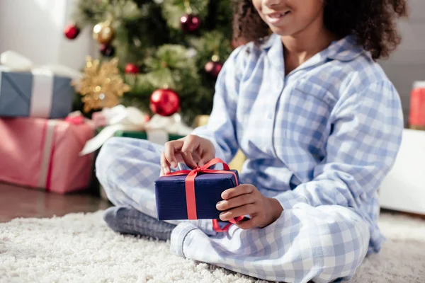 Zugeschnittenes Bild eines afrikanisch-amerikanischen Kindes im Pyjama, das zu Hause ein Weihnachtsgeschenk öffnet — Stockfoto