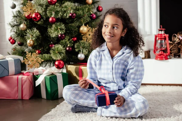 Niño afroamericano adorable feliz en pijama abriendo regalo de Navidad en casa y mirando hacia otro lado - foto de stock