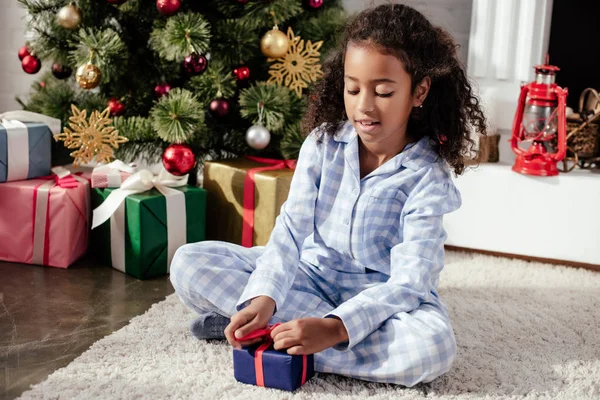 Adorable african american child in pajamas opening christmas gift at home — Stock Photo