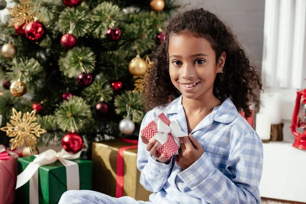Sonriente adorable afroamericano niño en pijama celebración de regalo de Navidad y mirando a la cámara en casa - foto de stock