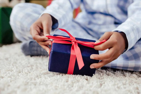 Imagen recortada de niño afroamericano en pijama abriendo regalo de Navidad en el suelo en casa - foto de stock
