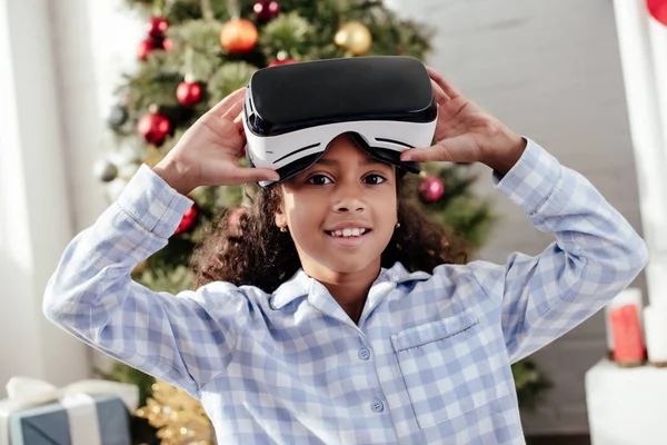 Niño afroamericano en pijama usando auriculares de realidad virtual y mirando a la cámara en casa, concepto de Navidad - foto de stock