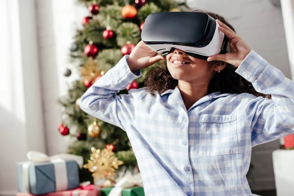 Cheerful african american child in pajamas using virtual reality headset at home, christmas concept — Stock Photo