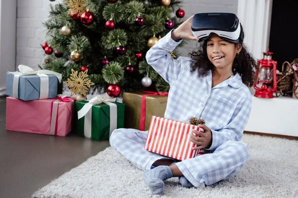 Niño afroamericano en pijama y casco de realidad virtual sacando la lengua y guiñando el ojo en casa, concepto de Navidad - foto de stock