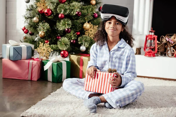 Niño afroamericano sonriente en pijamas y auriculares de realidad virtual que sostienen presente en casa, concepto de Navidad - foto de stock