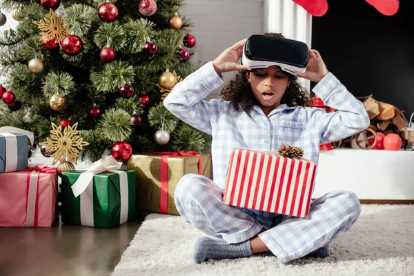 Surprised adorable african american child in pajamas and virtual reality headset looking at christmas gift at home — Stock Photo