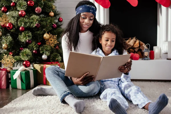 Cheerful african american mother and daughter reading book together at home, christmas concept — Stock Photo