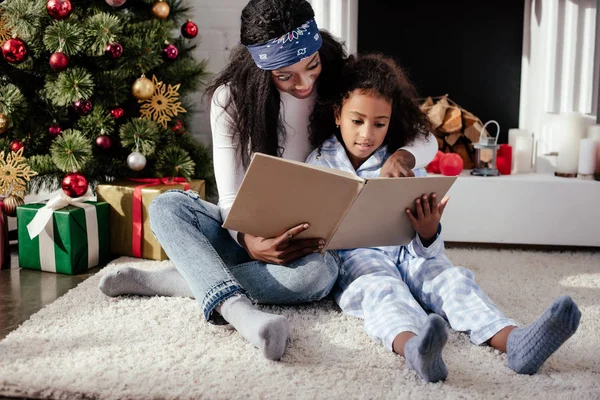 Madre afroamericana y su hija leyendo libro juntos cerca del árbol de Navidad en casa - foto de stock
