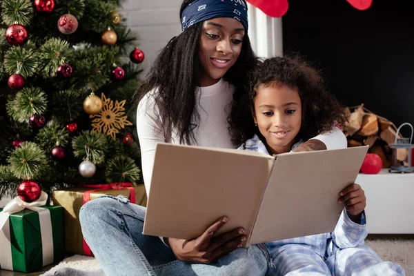 Sonriente afroamericano madre e hija leyendo libro juntos en casa, concepto de Navidad - foto de stock