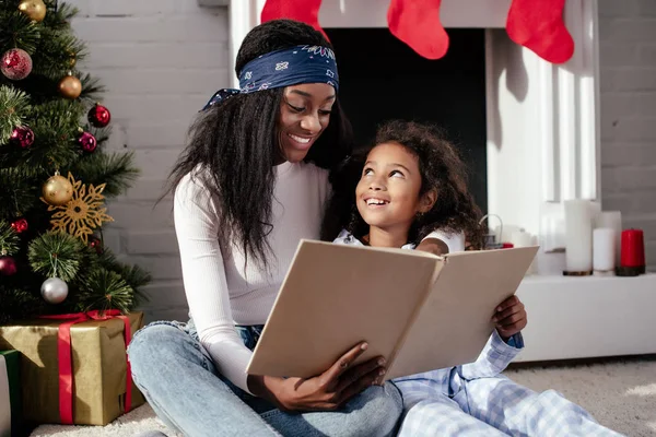 Happy african american mother and daughter reading book together at home, christmas concept — Stock Photo
