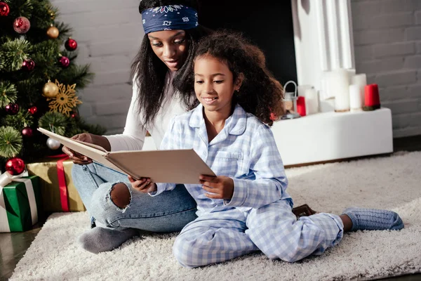 Madre afroamericana y su hija leyendo libro en el suelo en casa, concepto de Navidad - foto de stock