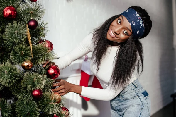 Belle femme afro-américaine décorer arbre de Noël avec des boules à la maison et en regardant la caméra — Photo de stock